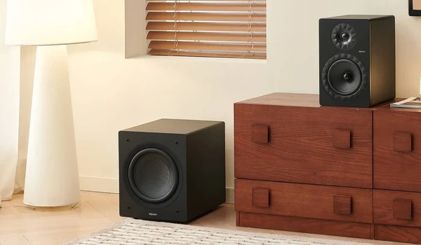 Elipson Classic Sub 8 posé au sol près d’une commode en bois, à côté d’une enceinte bibliothèque et d’un lampadaire