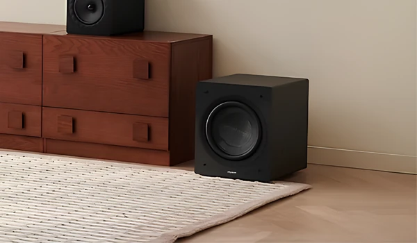 Elipson Classic Sub 8 noir posé au sol contre un mur, à côté d’un meuble en bois et d’un tapis, vue trois quarts