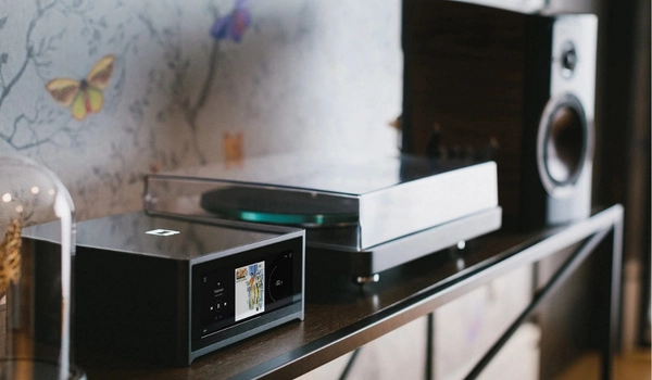 NAD M10 V3 sur une étagère en bois, à côté d'une platine vinyle sous capot et d'une enceinte bibliothèque en arrière-plan.