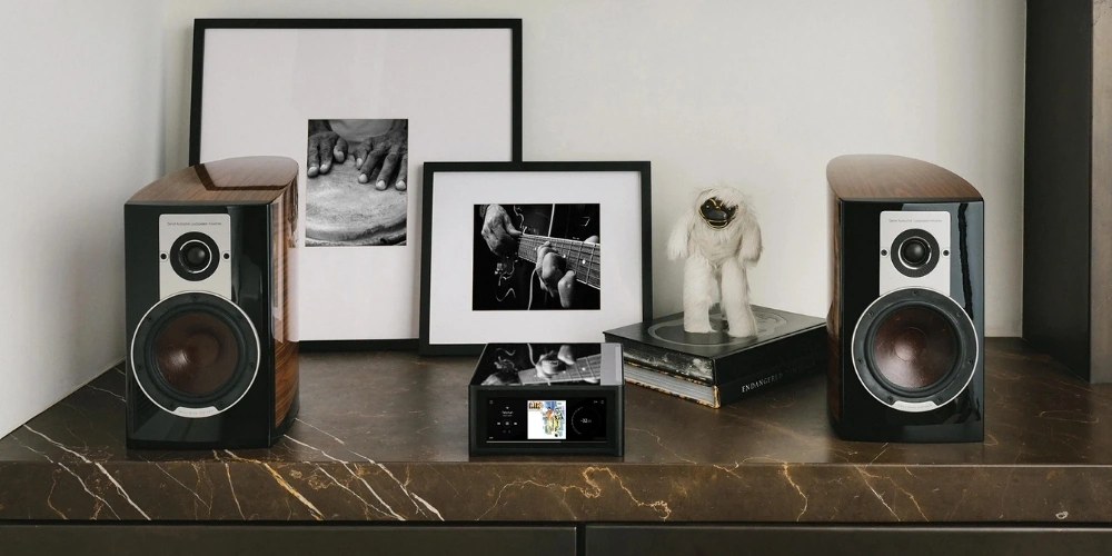NAD M10 V3 centré entre deux enceintes bibliothèque en bois sur une console en marbre, dans un salon avec cadres photo.