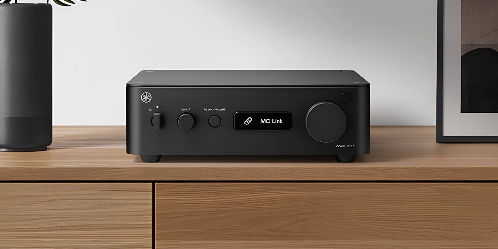 Yamaha WXC-70A en finition noire sur un meuble bois, écran MC Link allumé, entre une enceinte connectée et un cadre.