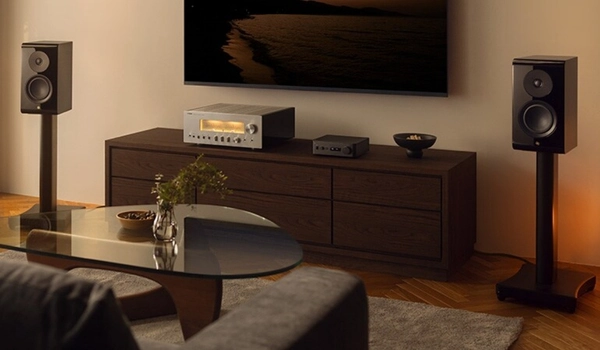 Salon avec ampli argenté et Yamaha WXC-70A sur un meuble bas, deux enceintes sur pieds et une TV murale allumée.